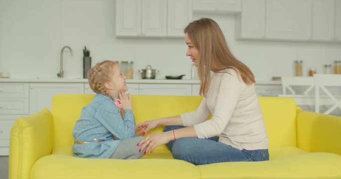 Positive excited little girl with pigtails and charming mother enjoying leisure together on sofa. Stunning mom and cheerful smiling elementary age girl communicating and bonding at home during weekend