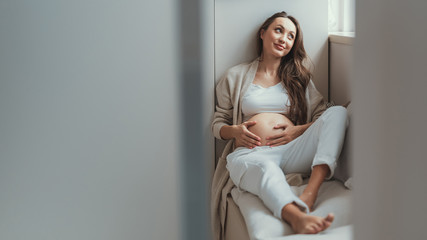 Calm mother looking aside and holding her tender hand on tummy