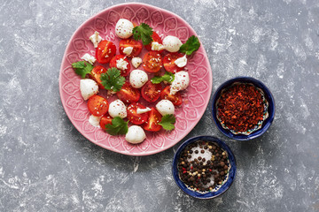 Caprese salad, cherry tomatoes, mozzarella and greens in a pink plate on a gray background. Italian food. Top view, flat lay.