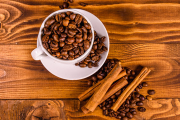 White cup filled with coffee beans and cinnamon sticks on wooden table. Top view