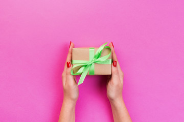 Woman hands give wrapped valentine or other holiday handmade present in paper with green ribbon. Present box, decoration of gift on pink table, top view with copy space