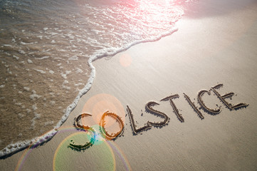 Solstice message handwritten in smooth sand with the light of the sun casting lens flare above an incoming wave on the beach