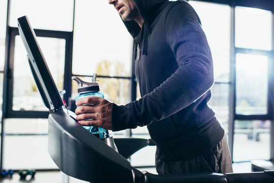 cropped image of sportsman in hoodie exercising on treadmill and holding sport bottle in gym