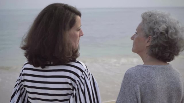 Mature brunette woman narrating something to senior woman on seashore. Two smiling ladies wearing casual clothing standing on sandy beach. Communication concept