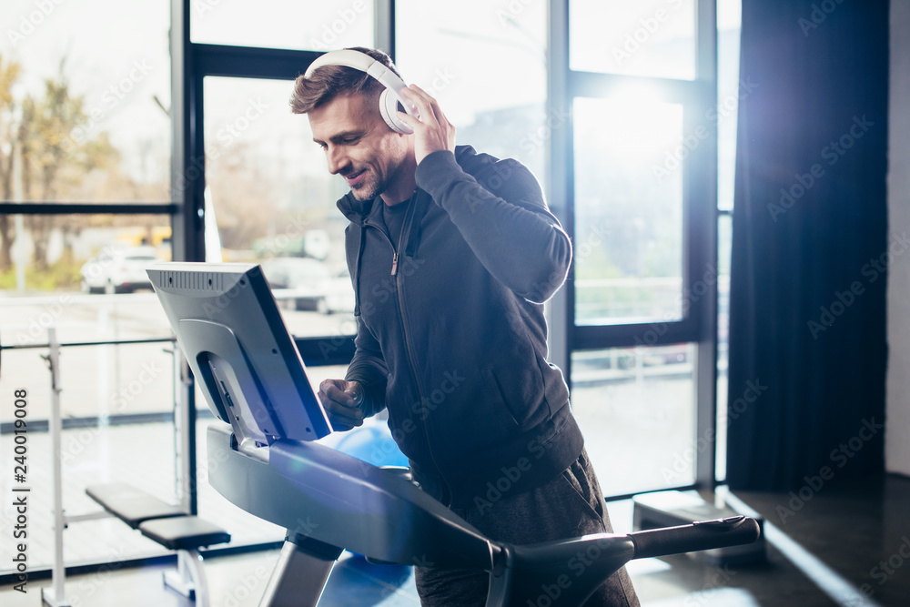 Wall mural handsome sportsman in hoodie exercising on treadmill and listening to music in gym - Wall murals