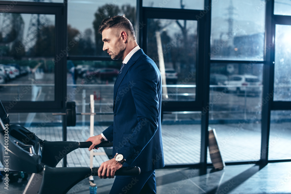 Wall mural side view of handsome businessman in suit exercising on treadmill in gym