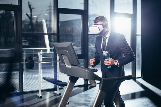 businessman in suit and virtual reality headset exercising on treadmill and holding sport bottle in gym