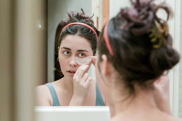 attractive young girl takes care of her skin by applying eye patches standing in front of mirror