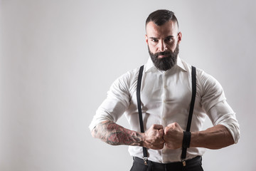 Uomo muscoloso tatuato  con camicia bianca e bretelle chiude i pugni deciso su sfondo bianco
