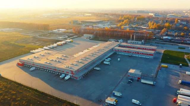 Aerial circling view of a logistics center with warehouse, loading hub with many semi-trailers trucks  load/unload goods at sunset
