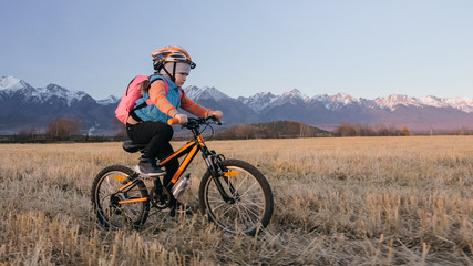 Obraz premium One caucasian children rides bike in wheat field. Little girl riding black orange cycle on background of beautiful snowy mountains. Biker motion ride with backpack, helmet. Mountain bike hardtail.