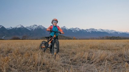 Obraz premium One caucasian children walk with bike in wheat field. Little girl walking black orange cycle on background of beautiful snowy mountains. Biker stand with backpack and helmet.