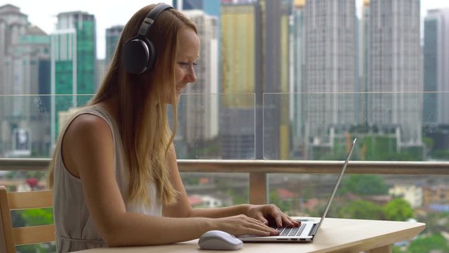 Young woman freelancer workes on her laptop at a balcony with a background of a city center full of skyscrapers. Remote techer has a conversation with a student through the internet
