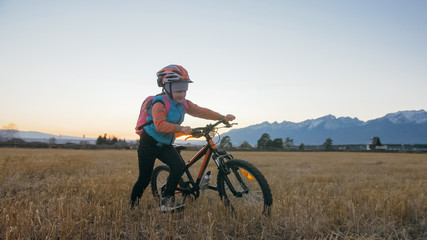 One caucasian children walk with bike in wheat field. Little girl walking black orange cycle on background of beautiful snowy mountains. Biker stand with backpack and helmet.