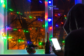 sad man in a hood with a smartphone in a blurred bokeh, on the background of the window decorated with garlands with an empty a bottle of champagne, in anticipation of the new year and Christmas