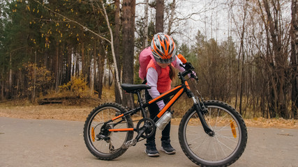 One caucasian children walk with bike in autumn park. Little girl walking black orange cycle in forest. Kid goes do bicycle sports. Biker motion ride with backpack and helmet. Mountain bike hard tail.