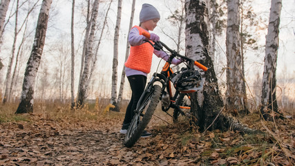Naklejka premium One caucasian children walk with bike in autumn park. Little girl walking black orange cycle in forest. Kid goes do bicycle sports. Biker motion ride with backpack and helmet. Mountain bike hardtail.