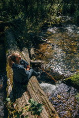 Beautiful girl sits on log and chilling after travel. Woman traveler in summer passes on wooden bridge in background of forest. Bridged lies on the river. Model of caucasian is chill and happy.