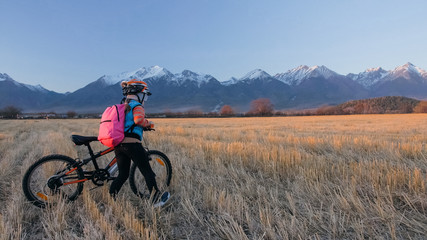 Obraz premium One caucasian children walk with bike in wheat field. Little girl walking black orange cycle on background of beautiful snowy mountains. Biker stand with backpack and helmet.