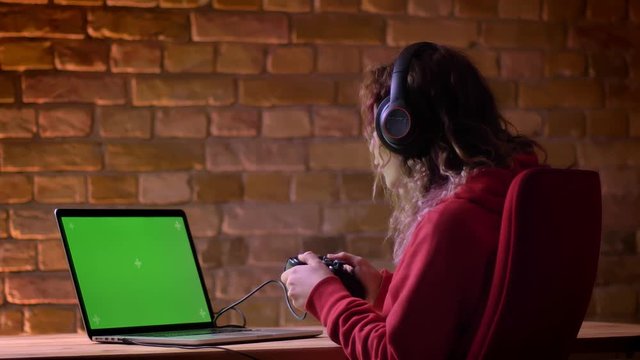 Rear view of young blogger in headphones happily playing video game using laptop and joystick on bricken wall background.