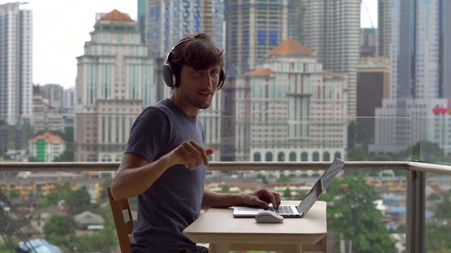 Young man freelancer workes on his laptop at a balcony with a background of a city center full of skyscrapers. Remote techer has a conversation with a student through the internet