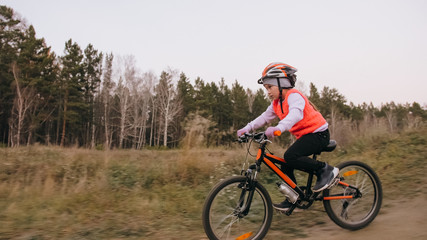 Fototapeta na wymiar One caucasian children rides bike road track in dirt park. Girl riding black orange cycle in racetrack. Kid goes do bicycle sports. Biker motion ride with backpack and helmet. Mountain bike hardtail.