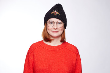 Adorable red headed young female in round spectacles, knitted winter outfit looks with pretty tender smile on freckled face, being glad  to meet such a good person as you, isolated on white wall.