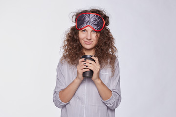 Pleased female glad to receive coffee at bed from lover, dressed in casual grey pajamas, has sleeping mask on head, has good sleep, isolated over white studio wall. Positive emotions early in morning.