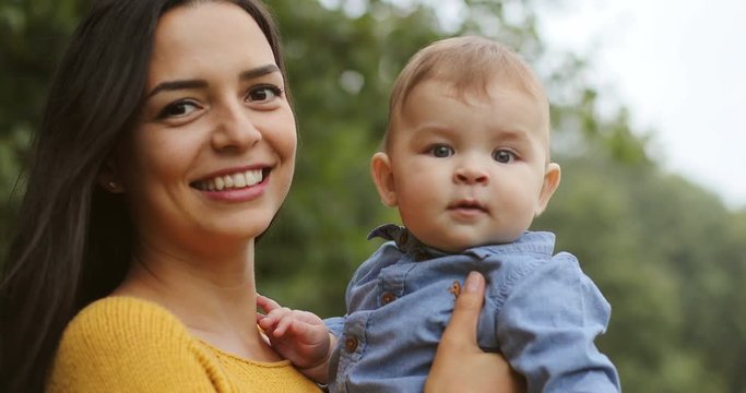 Close up of the cheerful and pretty young mother holding her cute toddler son and smiling to the camera in the summer at the park. Portrait. Indoor.