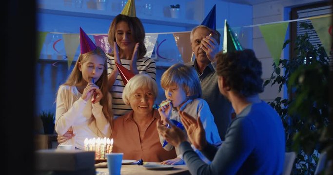 Happy Caucasian senior woman having her birhday party with a cake and being surounded by her grandchildren and daugther with husband. Inside.
