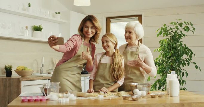 Caucasian happy and good looking mother standing at the kitchen with daughter and her grandmother and taking a selfie picture on the smartphone while making a dough. Inside.