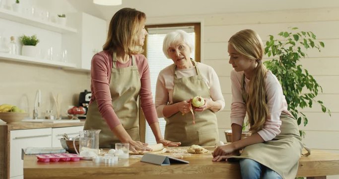 Small teenage girl sitting in the apron at the kitchen with a tablet computer and watching how her mother making a dough for a pie and grandmother peeling an apple. Indoors the kitchen.
