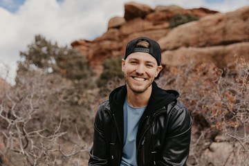 Portrait of Young Good Looking Casual Traveling Handsome Man Smiling Near Ancient Desert Red Rocks in Jacket Outside