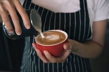Closeup image of female barista hands pouring milk and preparing fresh latte, coffee artist and preparation concept, red cup