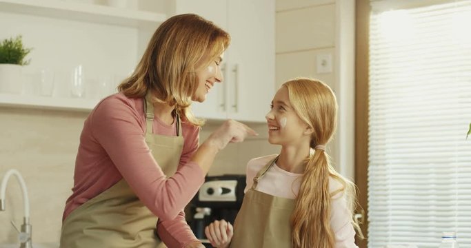 Portrait of the Caucasian happy mother and daughter smiling and laughing while putting flour on each other's noses while cooking together.