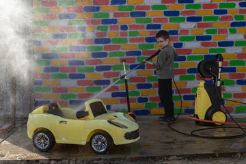 Happy child washes yellow toy car from high pressure washer in front of a multi-colored brick wall....