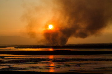 forest fire on the horizon with heavy dark smoke during the orange sunset with a frozen river on the foreground
