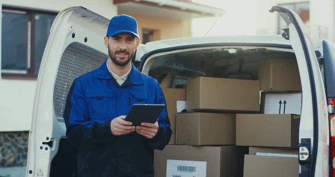 Portrait shot of the caucasian attractive deliveryman standing at the van full of parcels with a tablet computer in hands. Outside.