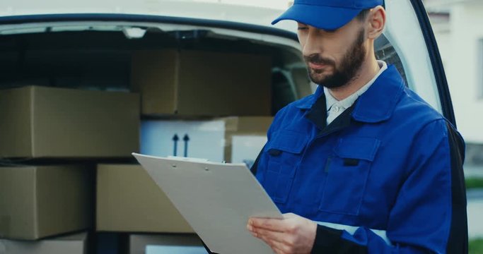 Close up of the Caucasian attractive male worker writing a document at the van full of carton parcels. Outside.