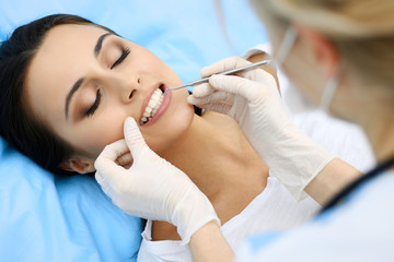 Naklejka premium Young female patient visiting dentist office.Beautiful woman with healthy straight white teeth sitting at dental chair with open mouth during oral checkup while doctor working at teeth