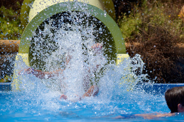 Caucasian boy gliding down slide in aqua park, making numerous spalshes around himself. He is...