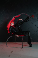 dangerous cruel fetish girl with red hair posing in black bdsm latex clothes at the chair from the...
