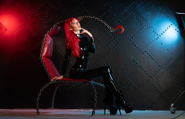 dangerous cruel fetish girl with red hair posing in black bdsm latex clothes at the chair from the...