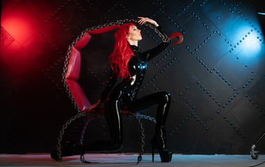 dangerous cruel fetish girl with red hair posing in black bdsm latex clothes at the chair from the...