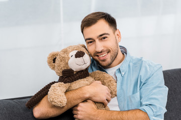 handsome man smiling, embracing teddy bear and looking at camera in apartment