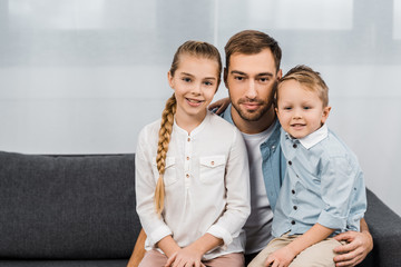 smiling father with cute children sitting on sofa and looking at camera in apartment