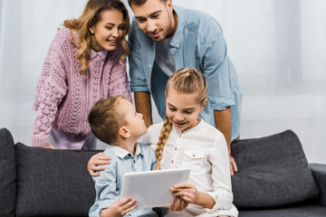 parents looking at cute siblings sitting on sofa and using digital tablet in living room