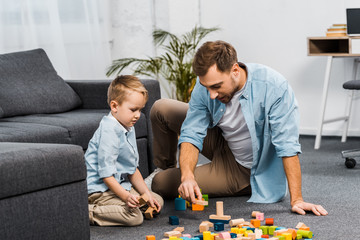 handsome man and cute boy playing with multicolored wooden blocks on floor in apartment