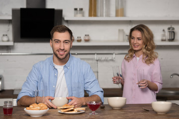 smiling man sitting at table and looking at camera with pretty wife holding glasses at background