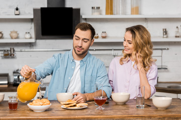 smiling woman looking at handsome husband pouring orange juice in glass on kitchen table